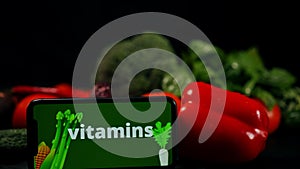 Vitamins ad on smartphone touchscreen app at background of healthful vegetables in darkness. Close-up raw organic