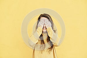 Vivacious laughing young woman covering her eyes with her hands on yellow background