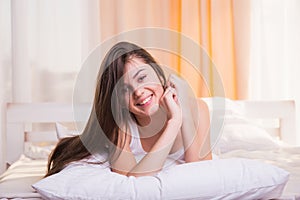 Vivacious young woman in bed lying on the bedclothes on her stomach with her chin on her hands