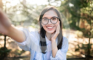 A vivacious young woman with glasses takes a selfie, her radiant smile and playful gesture