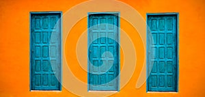 Vivid bright orange wall house facade with three blue-green closed doors on large empty orange wide wall texture background