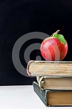 Vivid red apple with stem and green leaf on top of stack of old books on white desk in classroom. Blackboard. Back to school