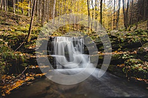 Vlasky River with waterfall illuminated with sun in an untouched landscape in the middle of an autumn mixed forest in red-orange