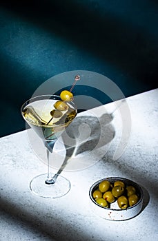 Vodka martini with olives, alcoholic cocktail drink with vodka and vermout, dark green background, bright hard light and shadow