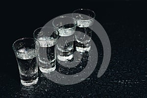 Vodka in shot glasses on black background, iced strong drink in misted glass.