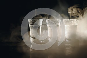 Vodka in shot glasses on black background, iced strong drink in misted glass.