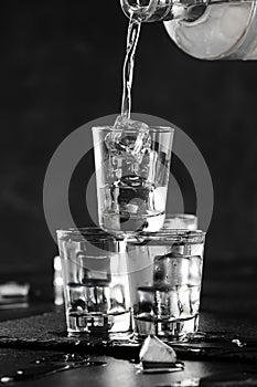 Vodka in shot glasses pouring out of the bottle on black stone background, iced strong drink in misted glass