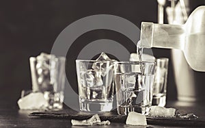 Vodka in shot glasses pouring out of the bottle on black stone background, iced strong drink in misted glass