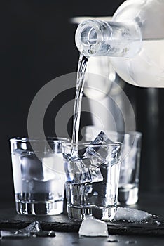 Vodka in shot glasses pouring out of the bottle on black stone background, iced strong drink in misted glass