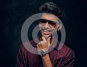 Vogue, fashion, style. Cheerful young Indian guy wearing a stylish shirt and sunglasses posing with hand on chin.