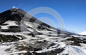 Volcanic landscape with dark cone of an active Asian volcano and white smoke against blue sky