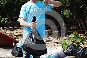 Volunteer in blue t-shirt holding garbage bag and giving thumbs down at rubbish dump, ecology man cleaning and picking up trash to