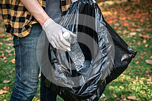 Volunteer man in gloves walking to carrying plastic black bag and collecting plastic bottles into bag