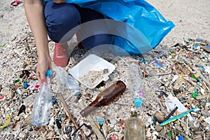 Volunteer tourist with hat is cleaning up garbage and plastic debris on dirty beach by collecting them into big blue bag