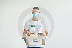 Volunteer work. Young arab man in volunteer t-shirt and medical mask, holding food donations box over light background