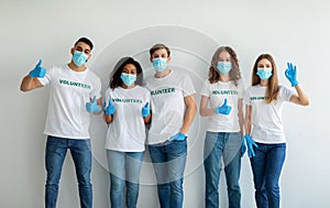Volunteering concept. Group of happy young diverse people in masks and gloves showing thumbs up, posing over light wall