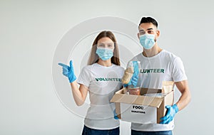 Volunteers in protective masks and gloves with food donations box over light background, woman pointing at free space