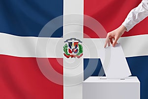 The voter throws the ballot into the box. Elections, Dominican Republic. Dominican Republic flag in the background