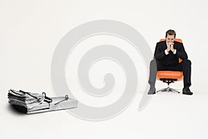 Wad of cash on mouse trap with pensive businessman on chair representing financial difficulties