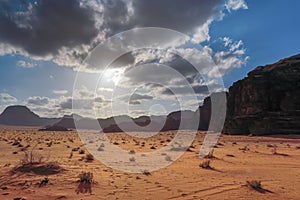 Wadi Rum desert, sun shines through cloudy sky over red dust and rocks, few small dry shrubs in foreground