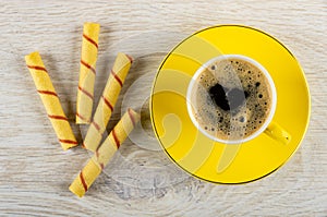 Wafer rolls, coffee in cup on yellow saucer on wooden table. Top view