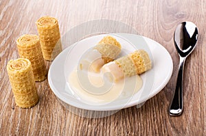 Wafer rolls with condensed milk in saucer, rolls, spoon on table