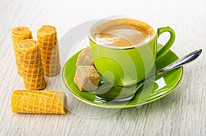 Wafer rolls, green cup with coffee, spoon, sugar on saucer on wooden table