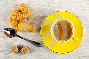 Wafer rolls, spoon, sugar, yellow cup with coffee on saucer on wooden table. Top view