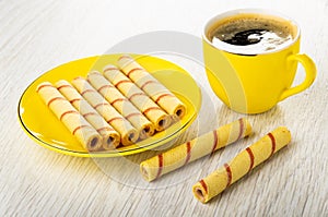 Wafer rolls in yellow saucer, cup of black coffee on wooden table