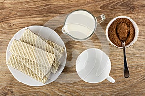 Wafers with porous chocolate in plate, pitcher with milk, cocoa with sugar, spoon in bowl, cup on wooden table. Top view