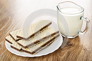 Wafers with porous chocolate in plate, transparent pitcher with milk on wooden table