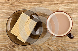 Wafers in saucer, brown cup of cocoa with milk on wooden table. Top view