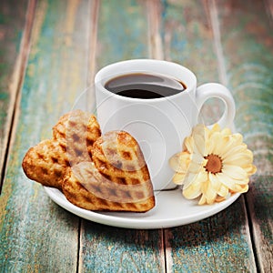 Waffle biscuits in shape of heart with cup of coffee and flower on vintage background