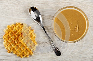 Waffle, spoon, bowl with peanut butter on wooden table. Top view