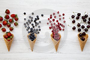 Waffle sweet ice cream cones with raspberries, cherries, strawberries and blueberries over white wooden background, overhead view