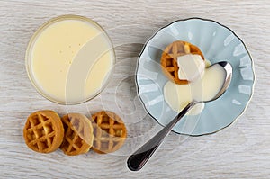 Waffles, teaspoon with condensed milk in saucer, bowl with condensed milk, waffles on table. Top view