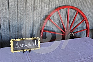 A wagon wheel leaning against the wall of a barn with a sign on a table for a traditional old-fashioned wedding celebration