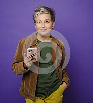 Waist up of attractive mature woman with smile holding smartphone and bank card in one hand isolated on colored studio background