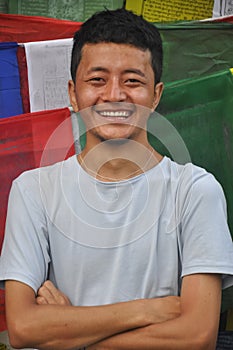 Waist up of a happy Ladakhi guy posing with crossed hands looking at camera, standing against Tibetan prayer flags