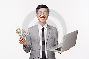 Waist-up of happy, pleased smiling young asian man in grey suit holding dollars, getting rich, using laptop as