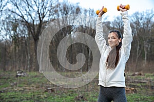 Waist up photo of a sporty brunette girl in white hoodie, white wirelless headphones who holds yellow dumbbells in her