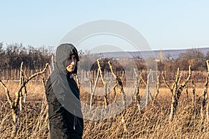 Waist up photo of the young serious man in black jacket stands among the field and looking at the camera, old garden