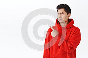 Waist-up portrait of asian angry man in red hoodie, frowning and grimacing, looking pointing judgemental, being photo