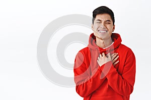 Waist-up portrait of charming asian man in red hoodie, hold hands on heart touched and thankful, smiling and laughing