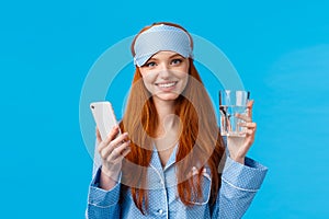 Waist-up portrait cheerful attractive glamour woman with red, ginger hair in sleep mask and pyjama, holding glass water