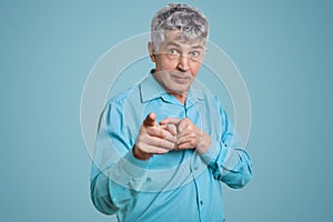 Waist up portrait of confident mature man pensioner points with both index fingers at camera, dressed in blue formal shirt, poses