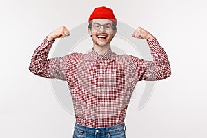 Waist-up portrait of encouraged and energized young confident slim guy in red beanie and glasses flex biceps, showing