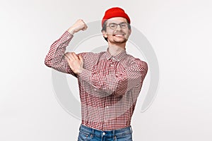 Waist-up portrait of funny adult slim guy in red beanie and glasses acting cool and tough, flex biceps showing how