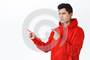 Waist-up portrait of handsome asian man looking judgemental or with hate, squinting frowning, pointing and staring with photo