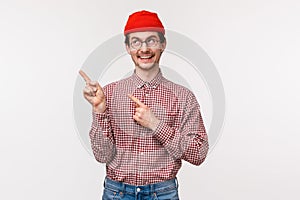 Waist-up portrait of happy smiling geeky guy in glasses and red beanie, pointing and looking upper left corner with